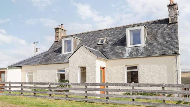 A two-story house with a sloped roof, a wooden fence in the foreground, and a clear sky in the background. - Loubcroy Farm House (Lairg)