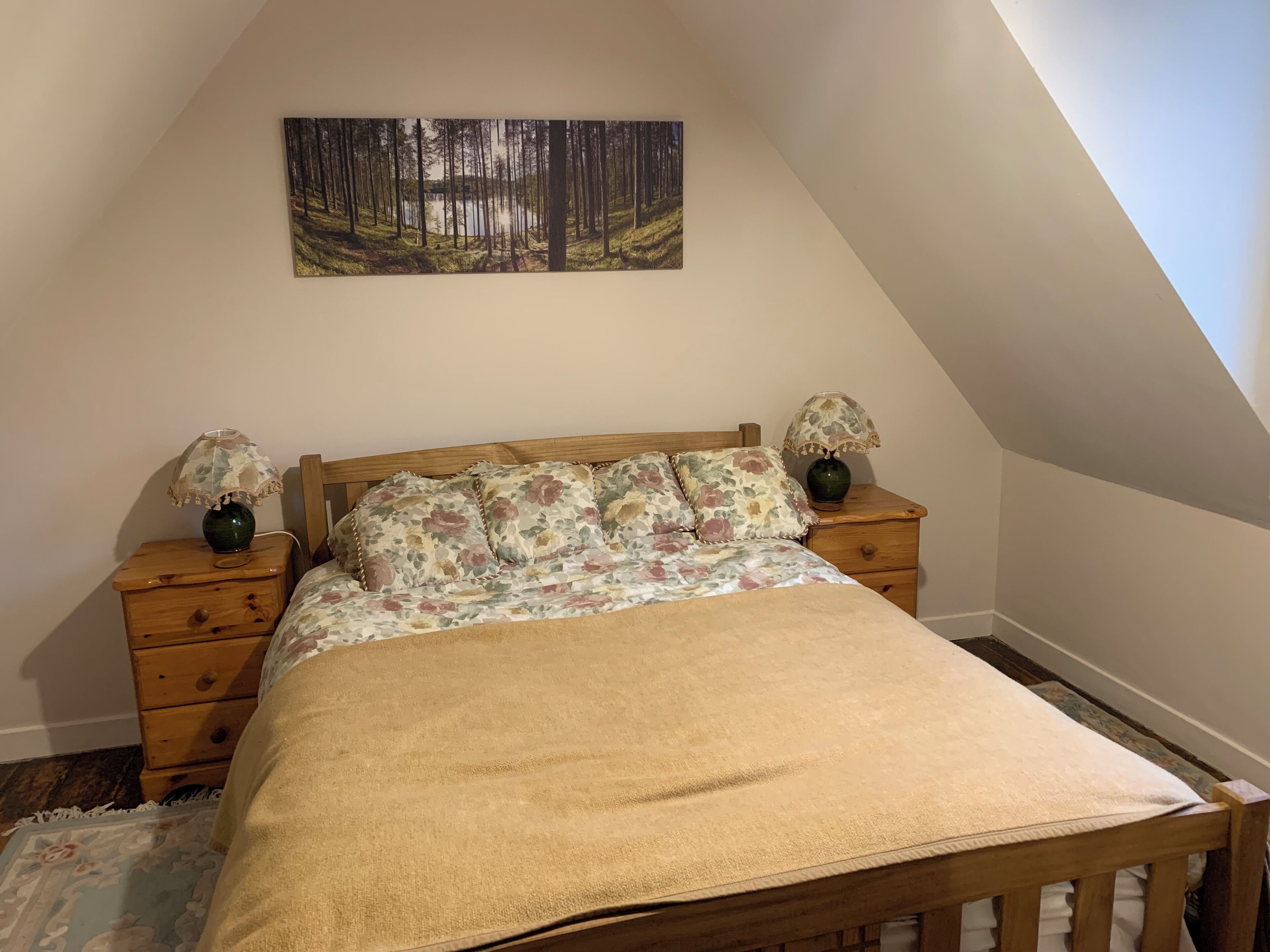 A bedroom with a wooden bed, floral bedding, two nightstands with lamps, a painting of a forest, and a sloped ceiling.