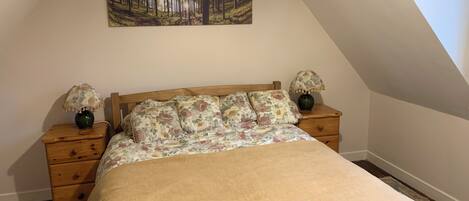 A bedroom with a wooden bed, floral bedding, two nightstands with lamps, a painting of a forest, and a sloped ceiling.