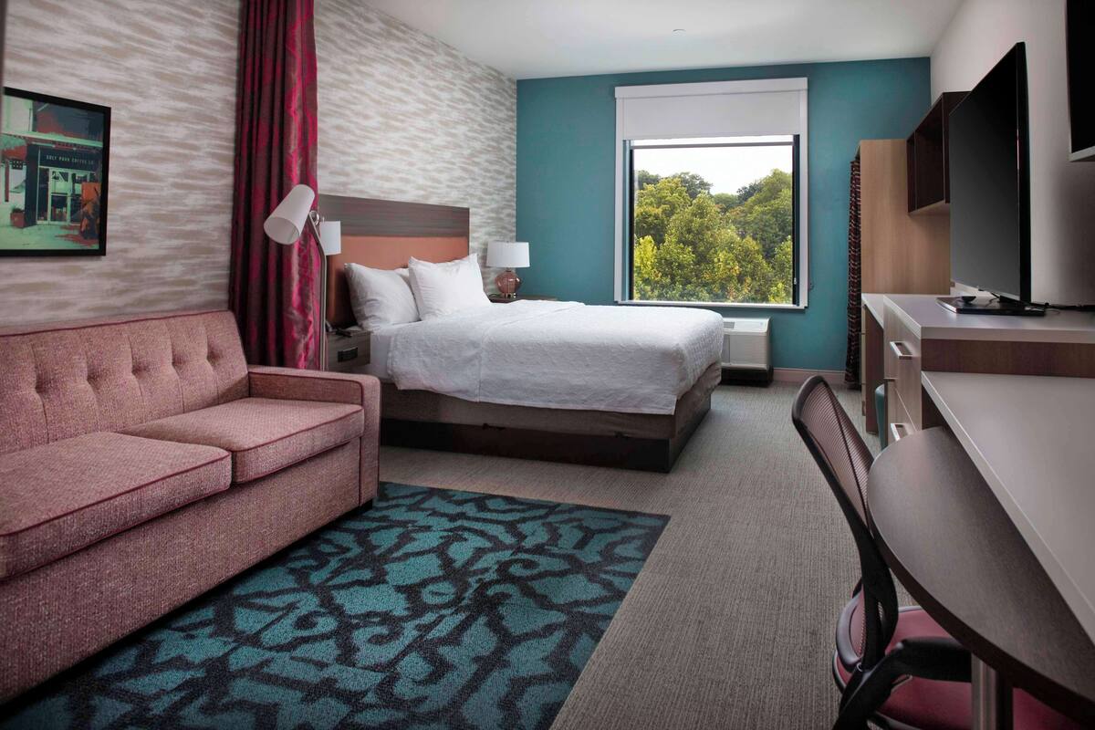 Een hotelkamer met een bed, een bureau, een bank en een groot raam met uitzicht op bomen.