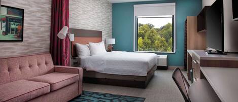 Habitación de hotel con cama, escritorio, sofá y una ventana grande con vistas a árboles.