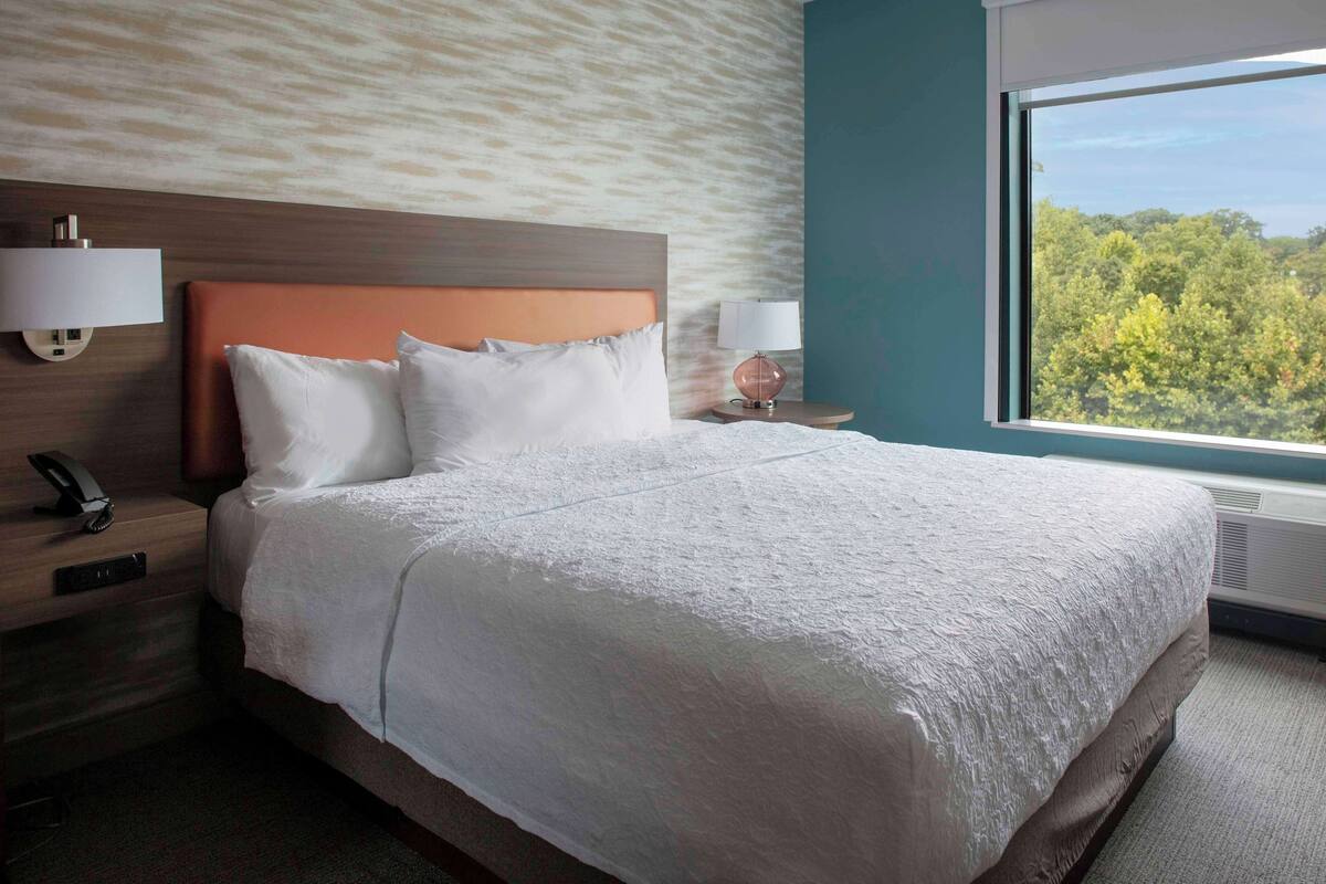 Een hotelkamer met een groot bed, twee nachtkastjes, een wandlamp en een raam met uitzicht op bomen.