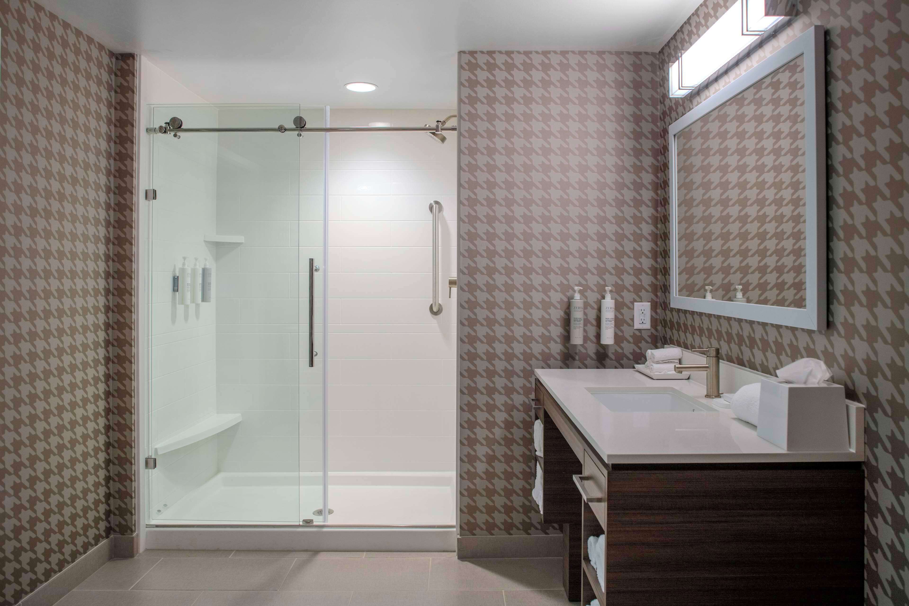 A modern bathroom with a glass-enclosed shower, a vanity with a mirror, and patterned wallpaper.