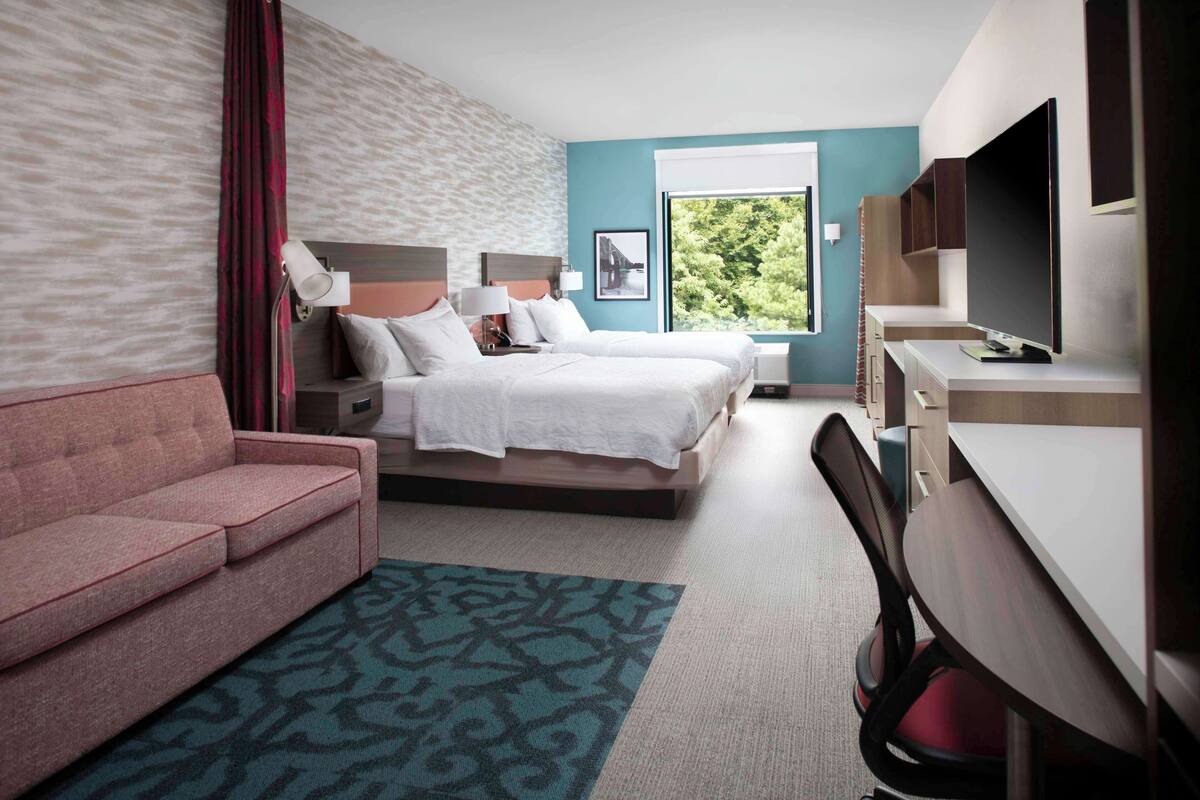 Een hotelkamer met een bed, een bureau, een stoel, een bank en een groot raam met uitzicht op groen.