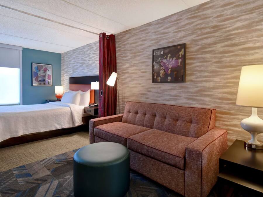 Quarto de hotel com cama, sofá, um banquinho redondo pequeno, mesa de cabeceira com abajur e parede com textura e um quadro emoldurado.