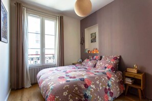 A bedroom with a floral bedspread, a bedside table with books, a wall-mounted lamp, and a window with curtains.