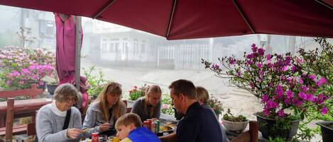A group of people dining at an outdoor table under a red umbrella, surrounded by potted plants and flowers.