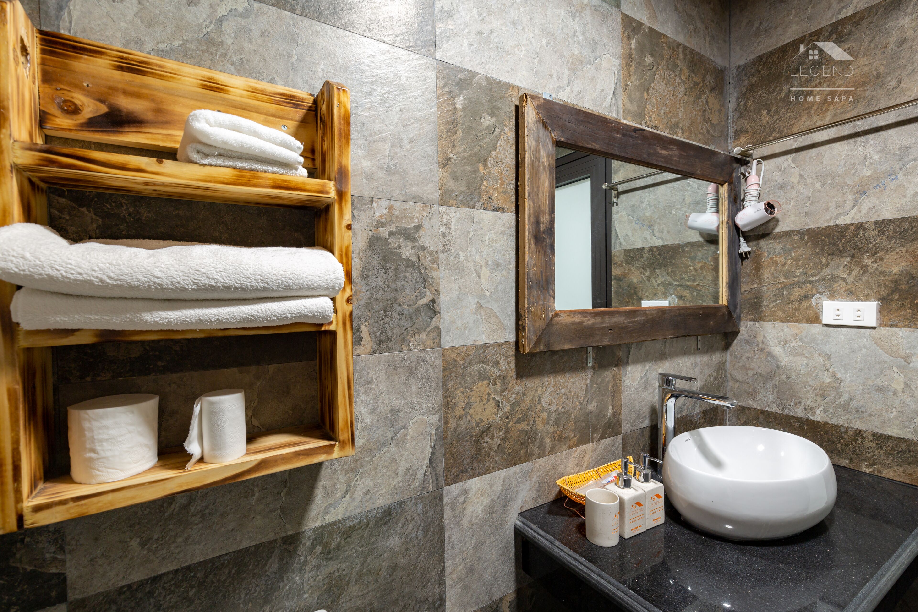 A bathroom with a wooden shelf holding towels, a sink, and toiletries.