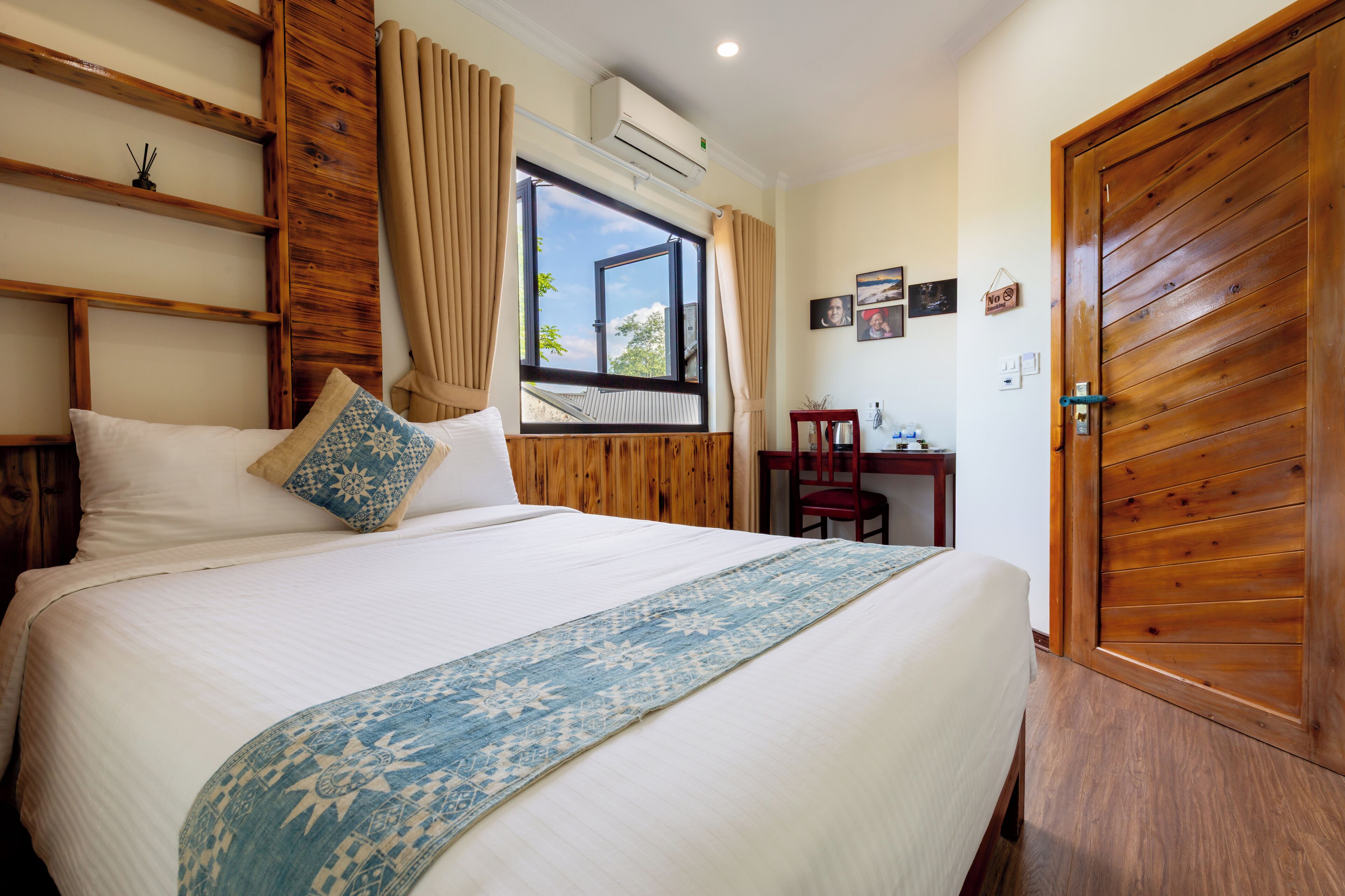 A bedroom with a large bed, a wooden wardrobe, a desk with a chair, and a window with curtains.