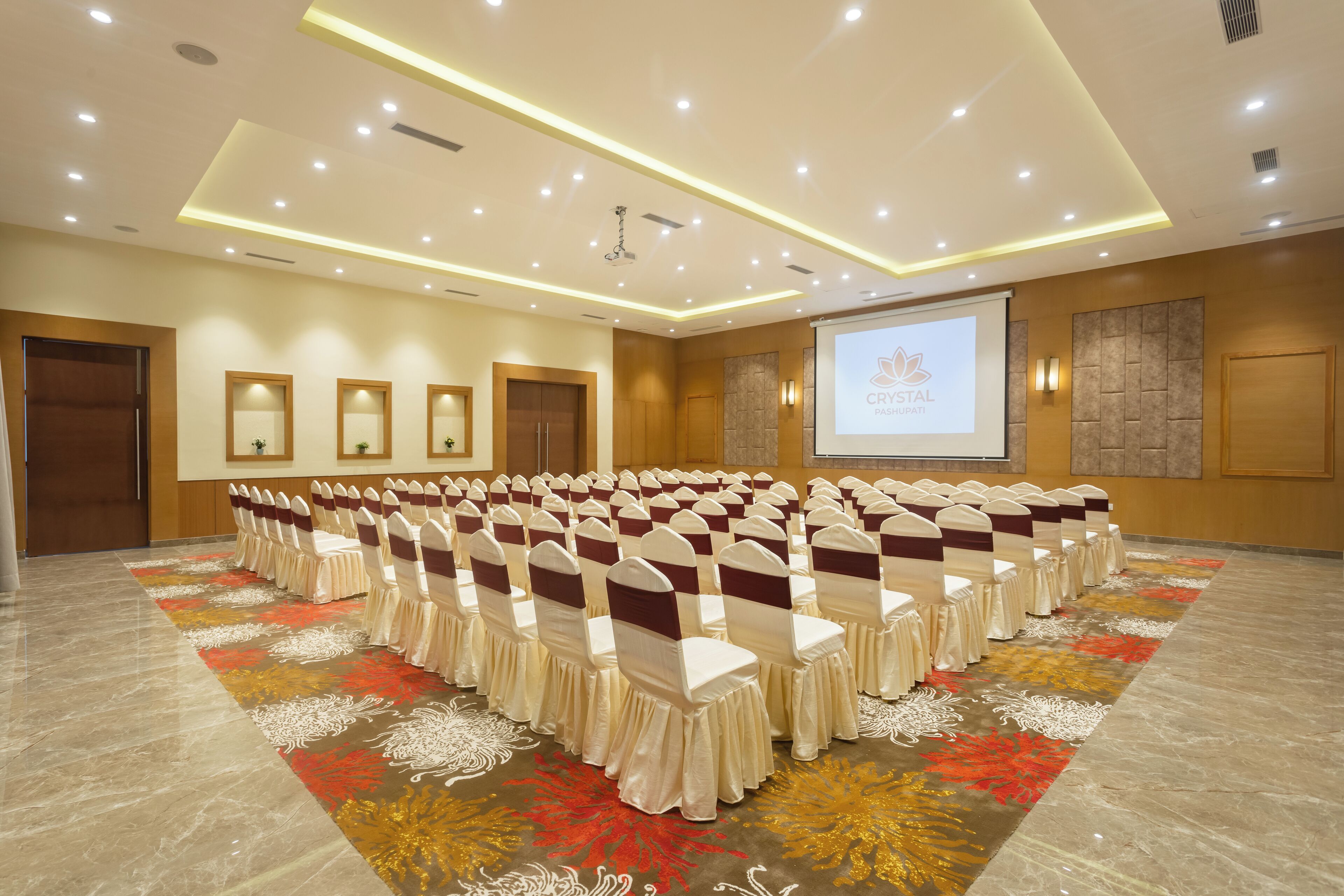 Un salón de conferencias con filas de sillas, una pantalla de proyector y una alfombra estampada con motivos florales.