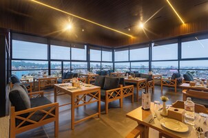 Un comedor moderno con mobiliario de madera, ventanas amplias que ofrecen vistas a la ciudad y un techo alto con iluminación lineal.