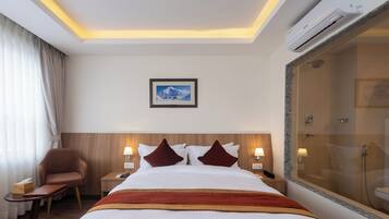 Una habitación de hotel con una cama, una silla, una mesita de noche con lámpara, un cuadro en la pared y un área de ducha.