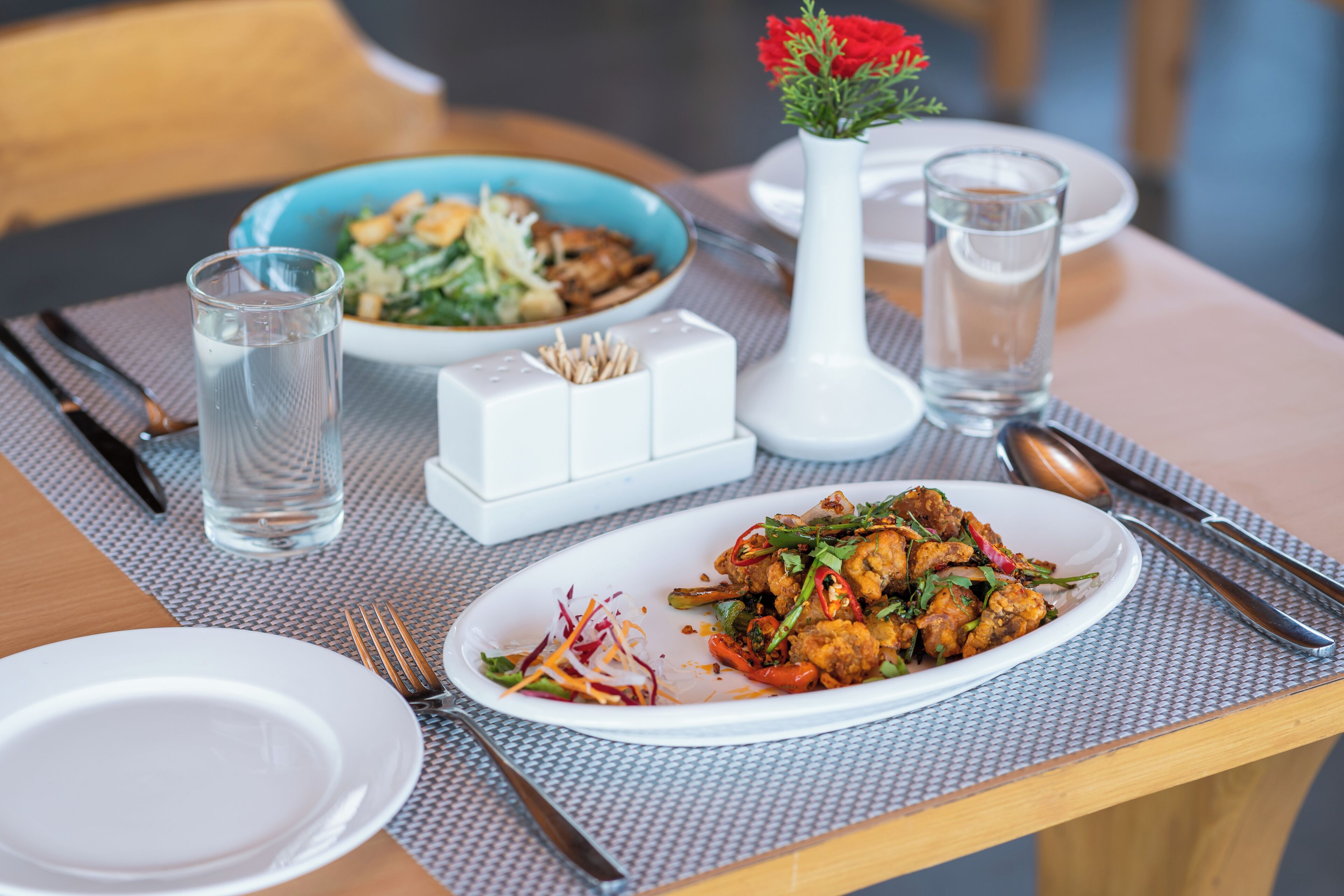 Una mesa de comedor dispuesta con platos, cubiertos, vasos y un florero con flores.