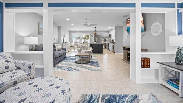 A modern living room with a sofa, armchair, and coffee table, a kitchen in the background, and a bookshelf with books. - Updated Waterfront Pool Home with Putting Green (Cocoa Beach)