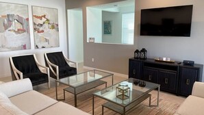A modern living room with a glass coffee table, two black chairs, a sofa, a flat-screen TV, and a dark wooden cabinet.