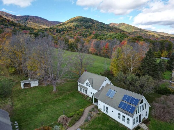 Une maison blanche avec un toit en pente et des panneaux solaires, entourée d’une pelouse verdoyante et d’arbres aux feuilles d’automne, le tout se détachant sur un paysage de collines.