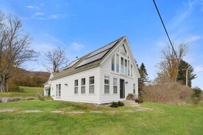 Une maison blanche à deux étages, avec un toit à pignon, de grandes fenêtres et une véranda à l’avant.