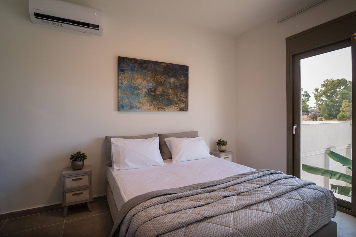 Un dormitorio con cama, una mesita de noche, una planta en maceta, un cuadro en la pared y un ventanal corredero que da a un balcón.