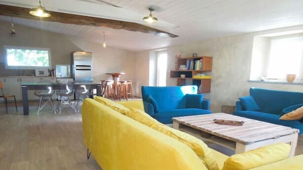 Un soggiorno con un divano giallo, poltrone blu e un tavolino in legno. C'è un'area pranzo con tavolo e sedie, e una cucina sullo sfondo.