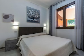 A bedroom with a bed, a nightstand, a wall-mounted painting, and a window with curtains.