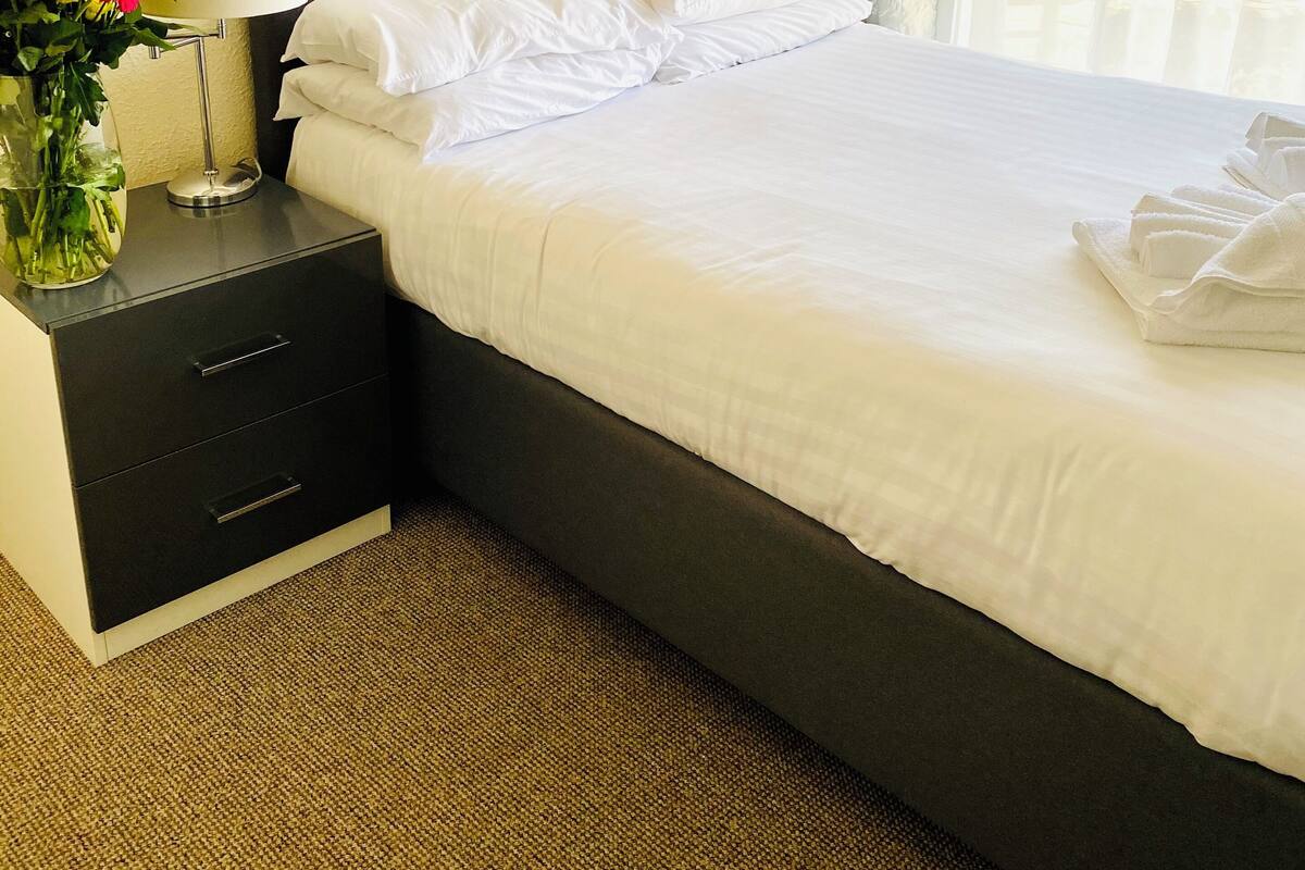 A neatly made bed with white linens, a grey upholstered headboard, and a bedside table with a lamp and a vase of flowers.