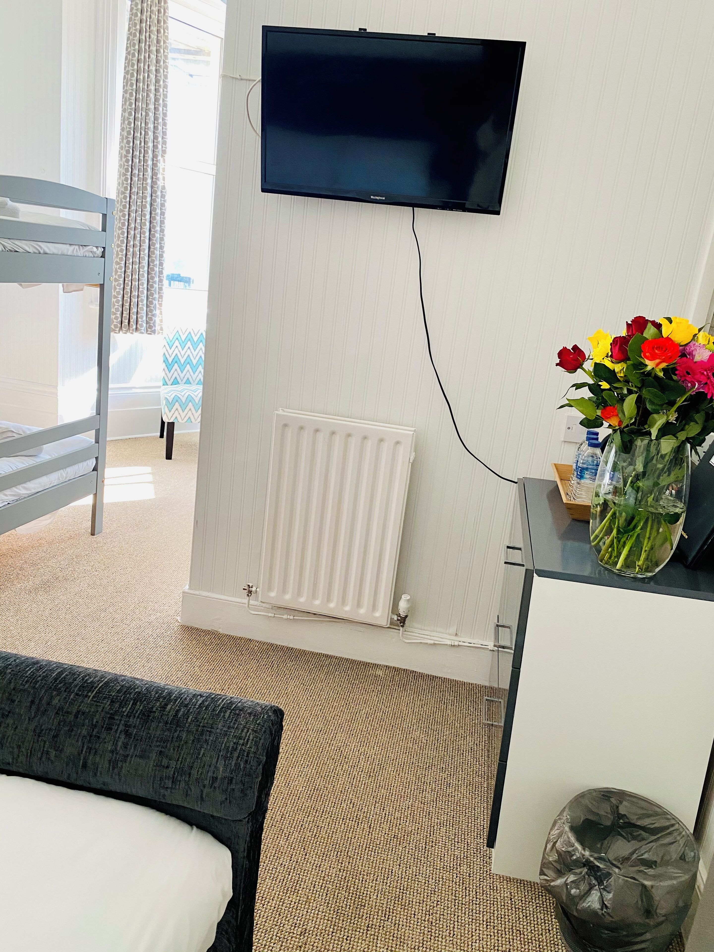 A room with a bunk bed, a flat-screen TV mounted on the wall, a radiator, a small table with a vase of flowers, and a trash bin.