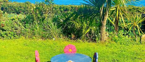 Una mesa redonda con sillas y respaldo con forma de flor, rodeada de abundante follaje y una palmera, con vista al océano en el fondo.