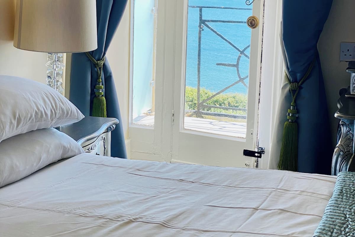 A bedroom with a bed, a nightstand, a lamp, and a view of the sea through a window with blue curtains.