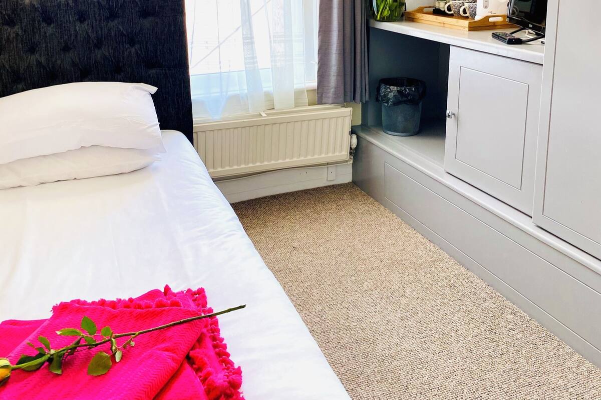 A hotel room with a bed, a bedside table with a coffee maker, a mirror, and a window with curtains.