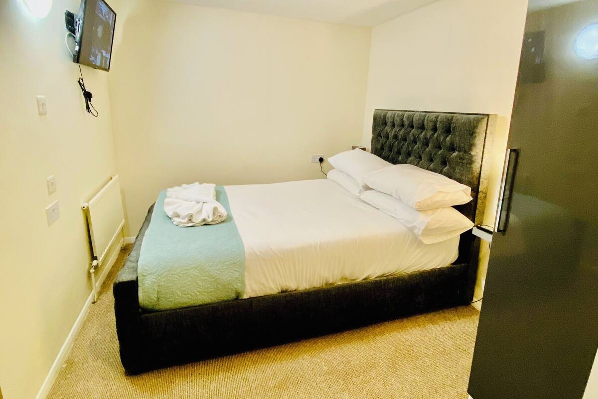 A small, neatly arranged hotel room with a single bed, a flat-screen TV mounted on the wall, and a refrigerator.