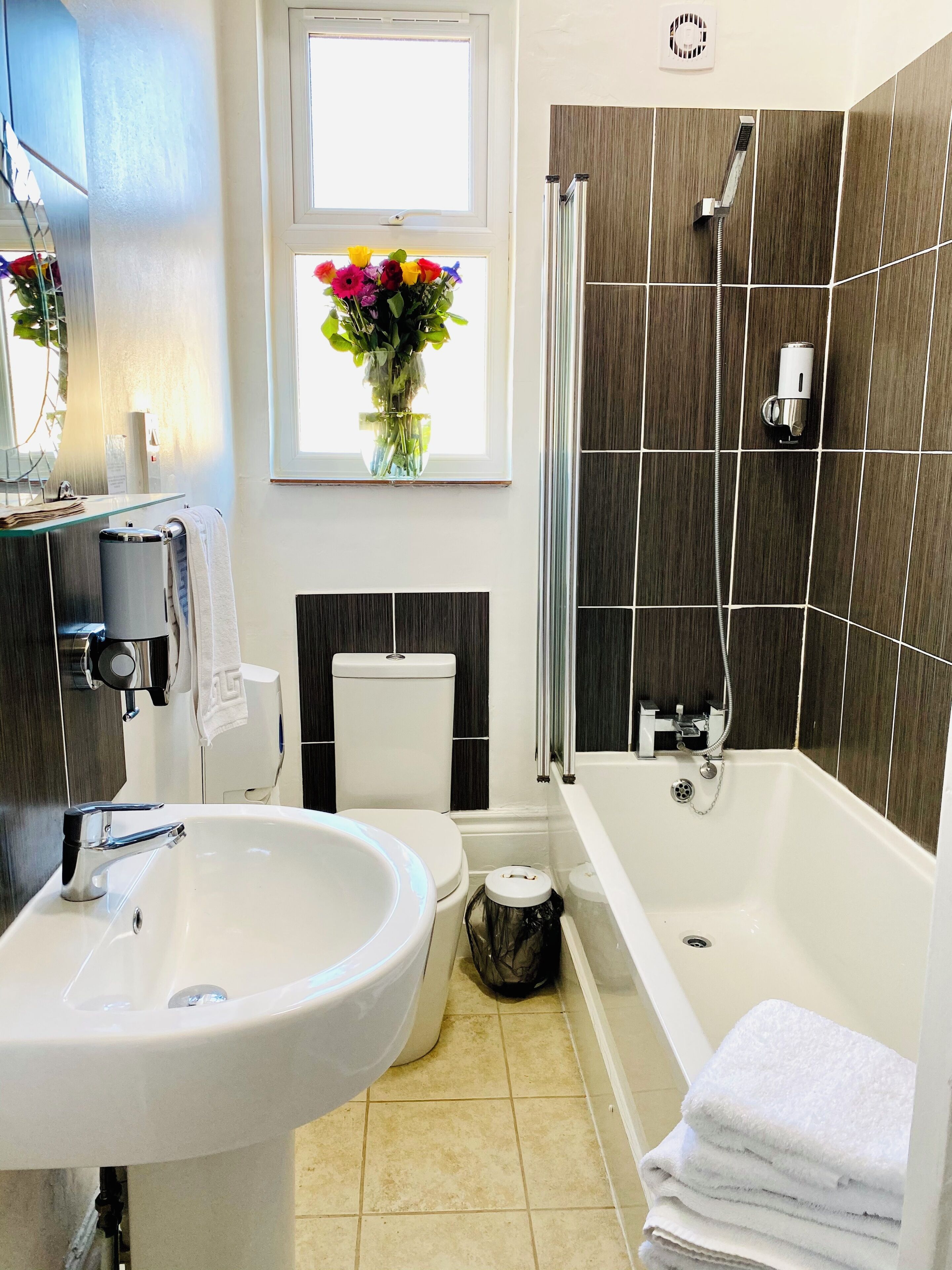 Un baño moderno con un lavabo, inodoro y bañera blancos. Se aprecia una ventana con un florero, y las paredes están cubiertas de azulejos.