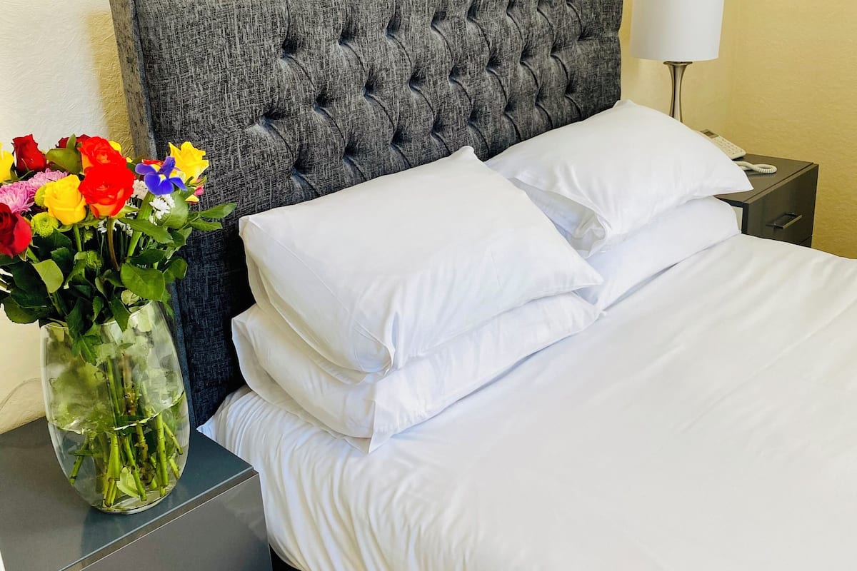 A neatly made bed with white linens, a grey tufted headboard, a bedside table with a lamp, and a vase with colorful flowers.