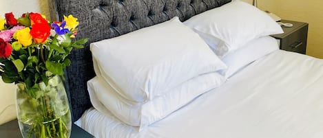 A neatly made bed with white linens, a grey tufted headboard, a bedside table with a lamp, and a vase with colorful flowers.
