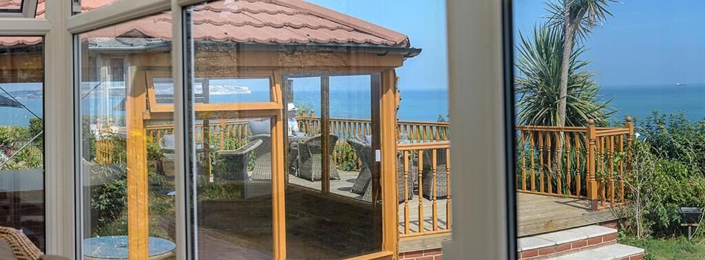 A glass-enclosed balcony with a wooden railing, overlooking a garden and the sea.
