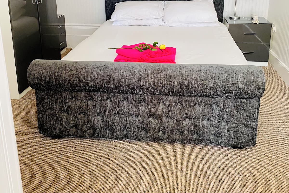 A neatly arranged hotel room with a bed, a grey upholstered bench, a nightstand with a lamp, and a wardrobe.