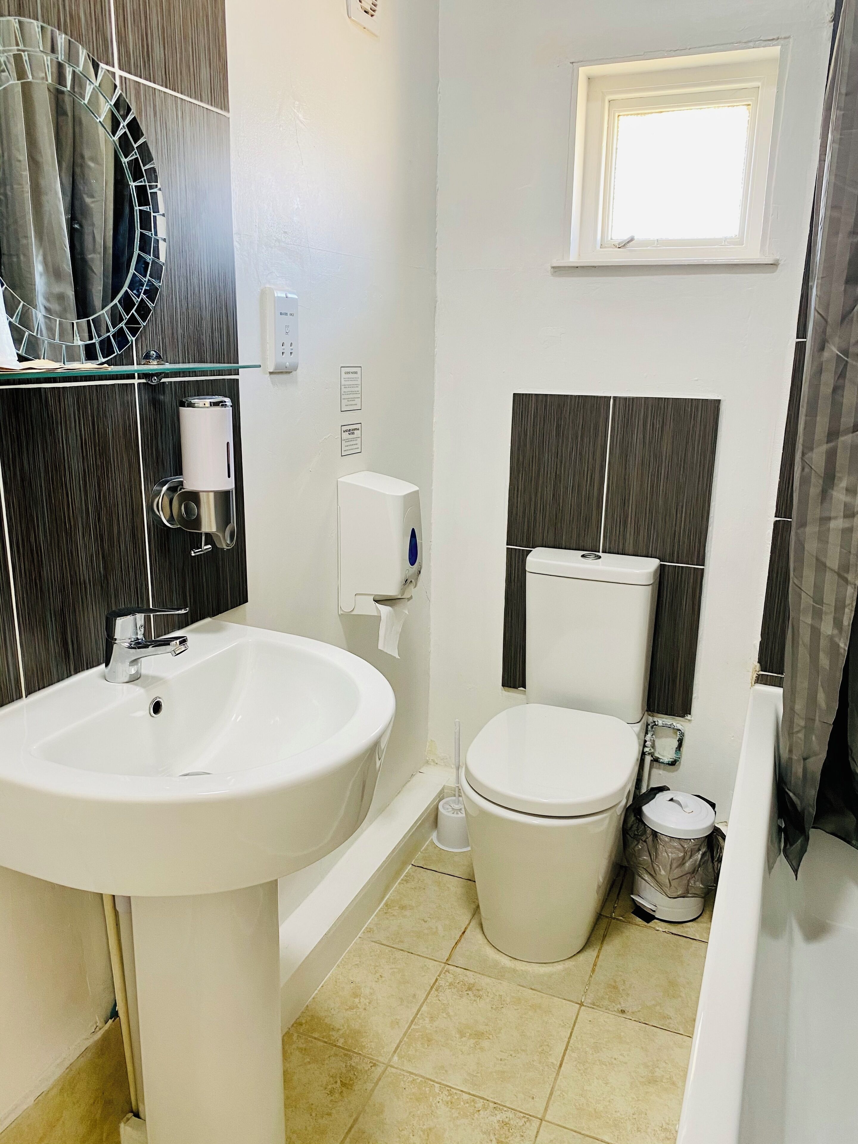 A bathroom with a white sink, toilet, and bathtub. There is a window above the toilet and a mirror with a circular frame on the wall.