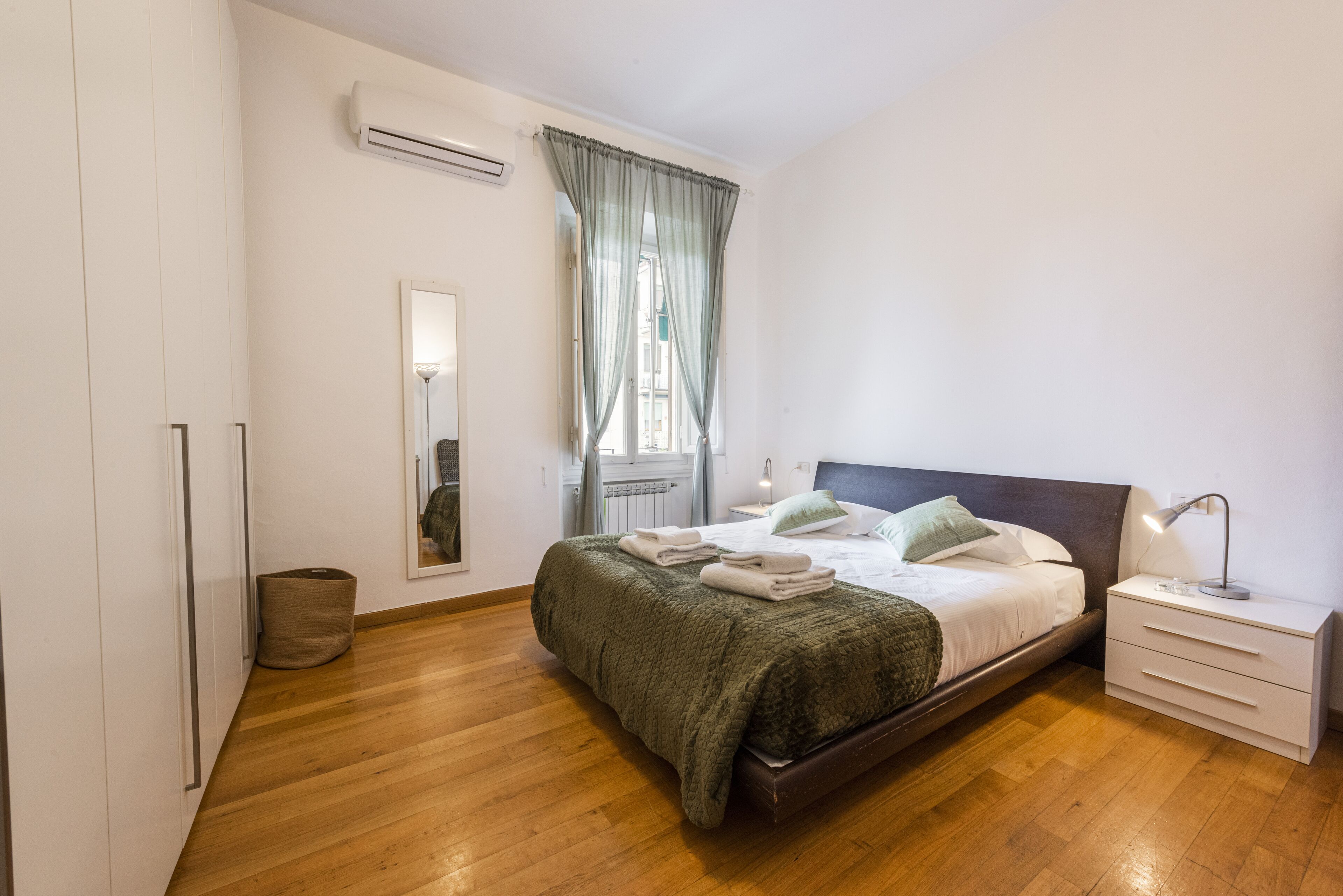 Une chambre avec un plancher en bois, un lit recouvert d’une couverture verte, une table de chevet avec une lampe, une fenêtre avec des rideaux et un appareil de climatisation.