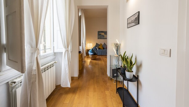 A hallway with a wooden floor, white walls, a radiator, a black chair, a potted plant, and a blue sofa in the background. - Casa Zamia by Mmega (Florence)