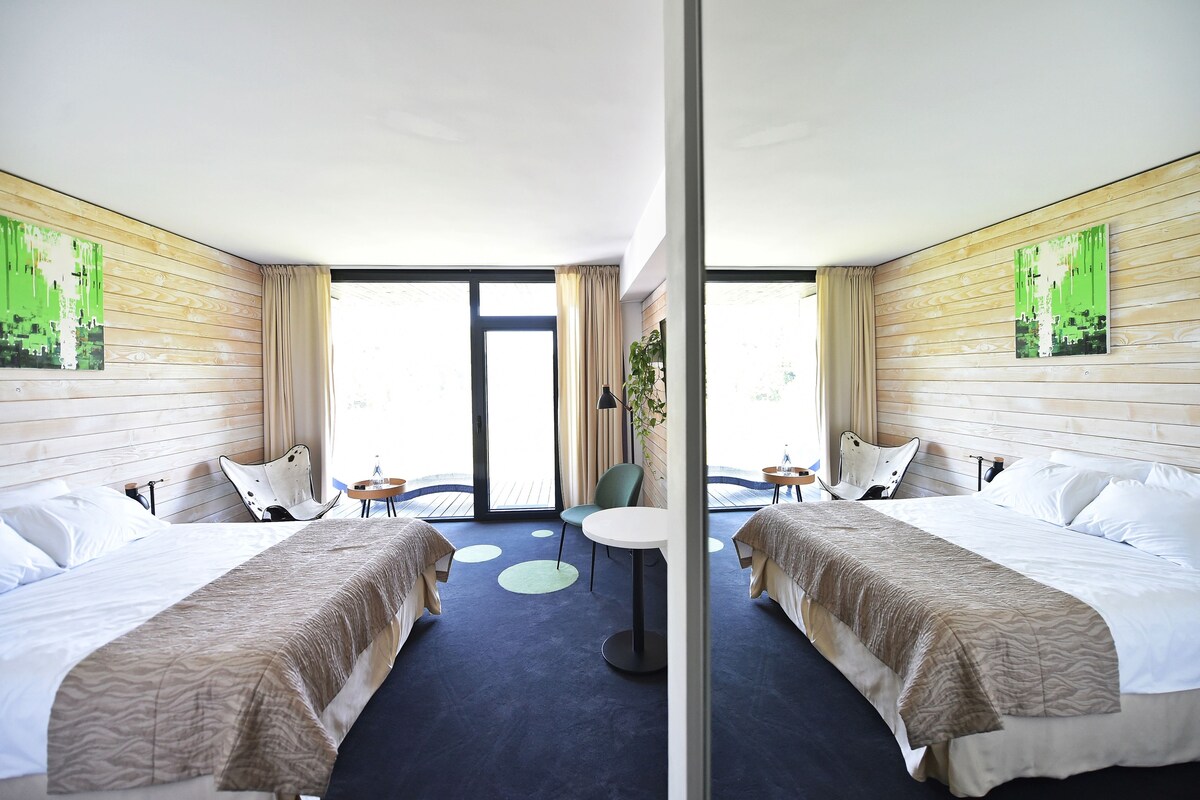 Habitación de hotel con dos camas, una mesa redonda pequeña y vistas al exterior a través de amplios ventanales.