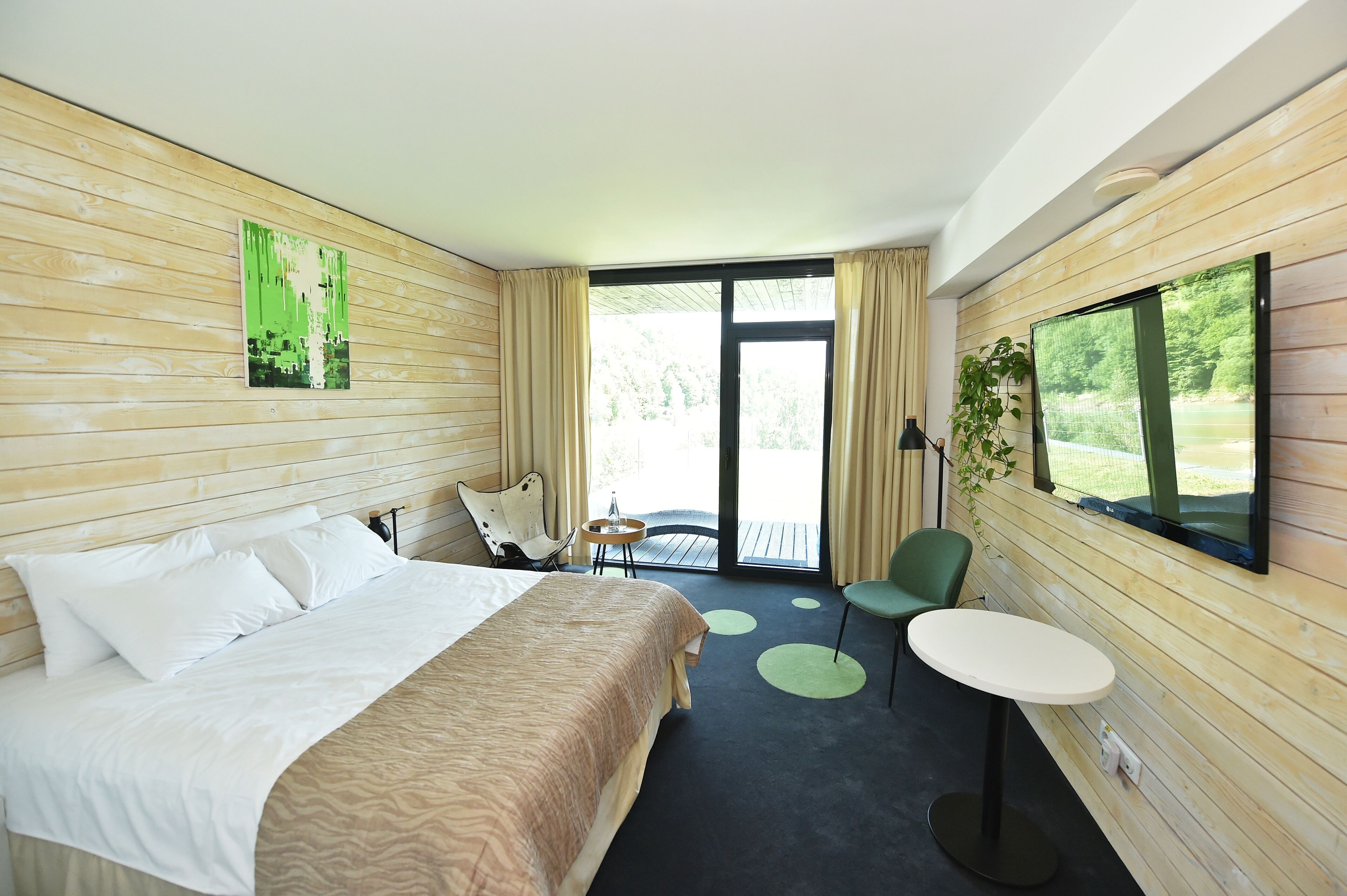 A hotel room with a bed, a small table, a chair, and a large window with a view of greenery.