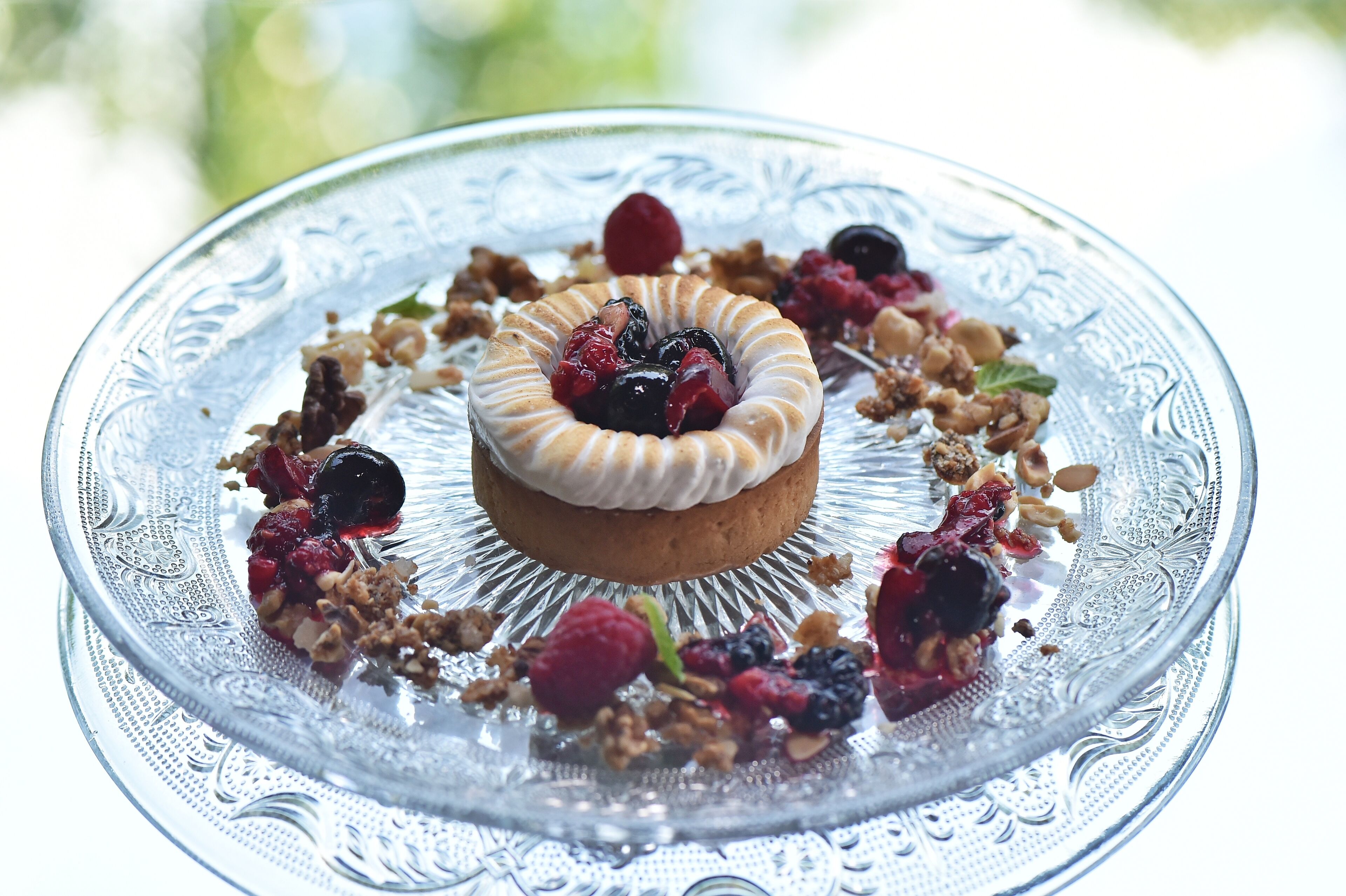 A dessert on an ornate glass plate with a central pastry, surrounded by berries and granola.