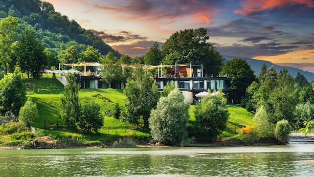 Un hotel moderno junto al lago, con un edificio grande de techo plano y numerosas ventanas, rodeado de abundante vegetación y un río.