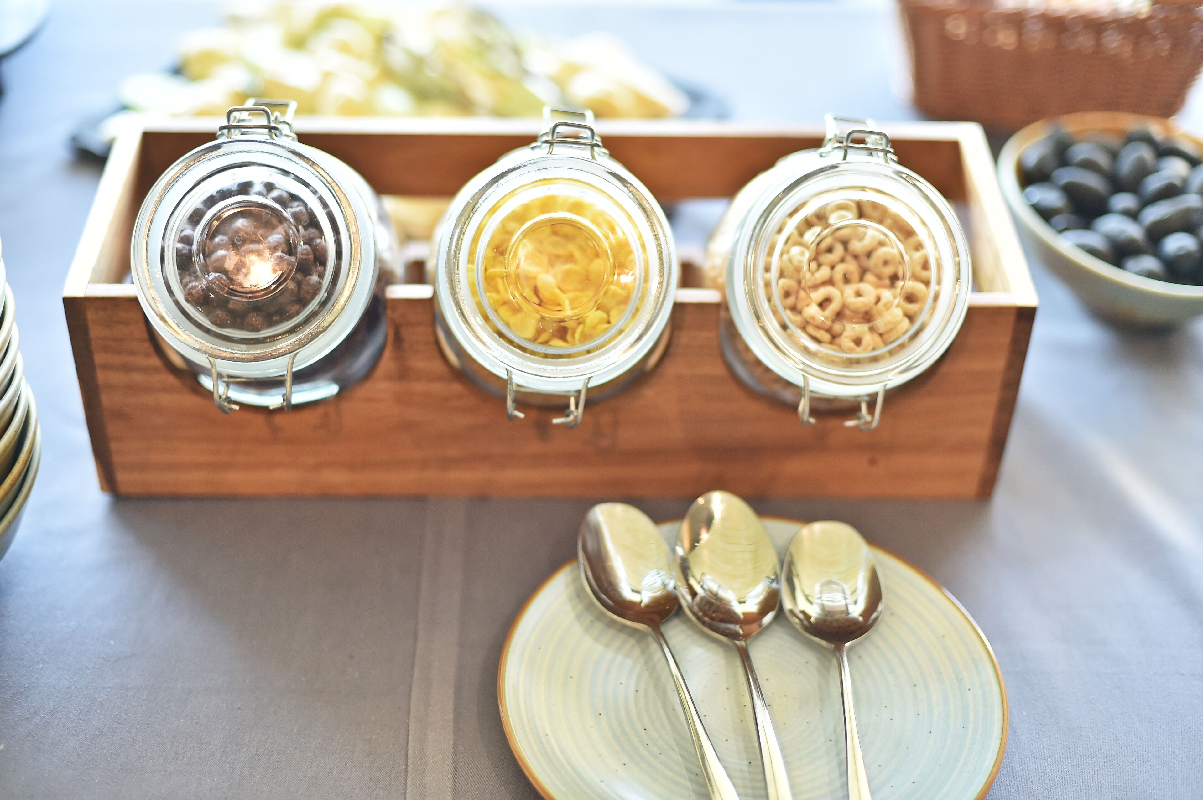 Una bandeja de madera con tres frascos de vidrio que contienen distintos alimentos, un cuenco de aceitunas y dos cucharas sobre un plato.