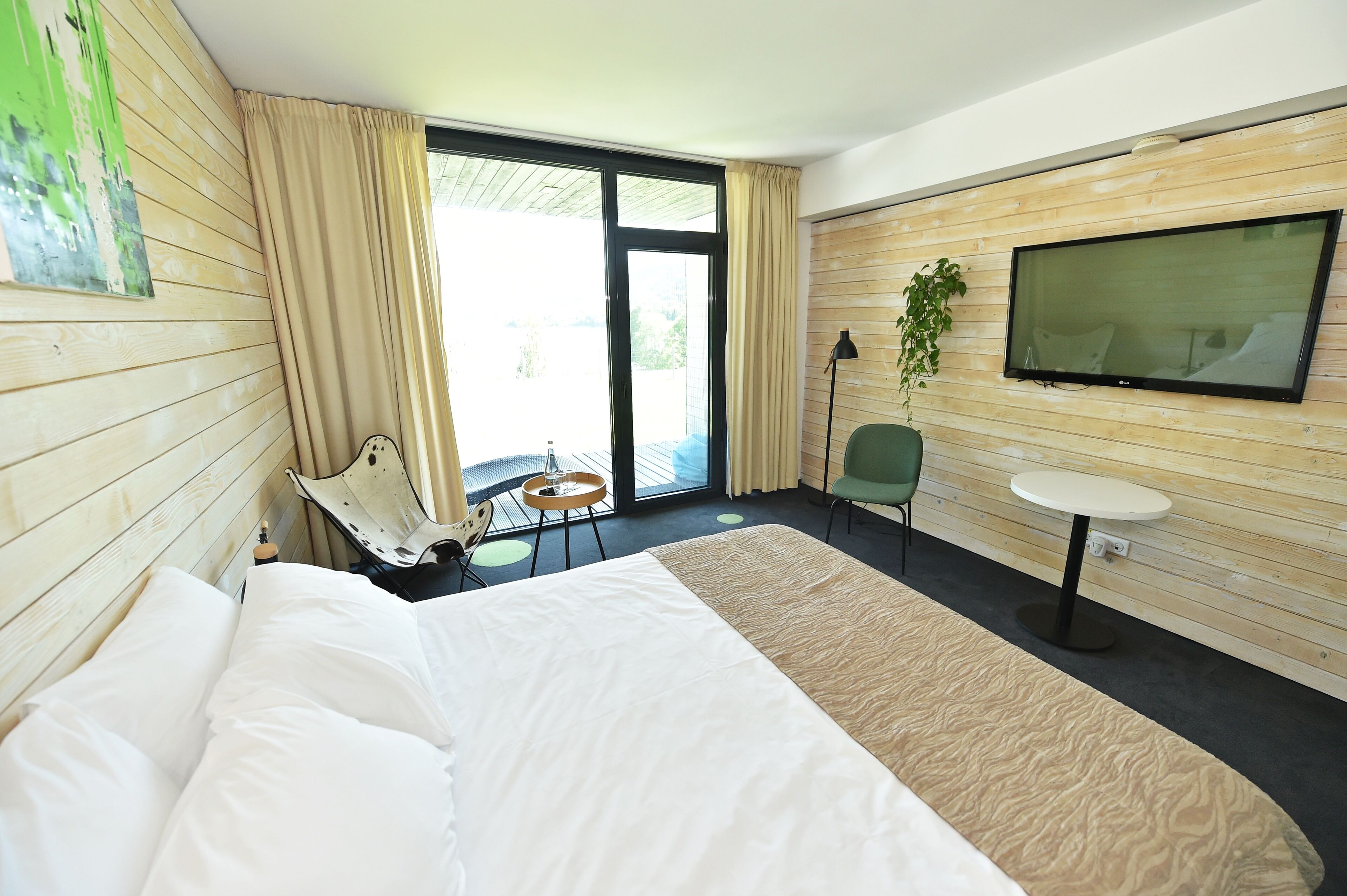 Une chambre d’hôtel avec un lit, une petite table, une chaise, un téléviseur et une grande fenêtre avec des rideaux.
