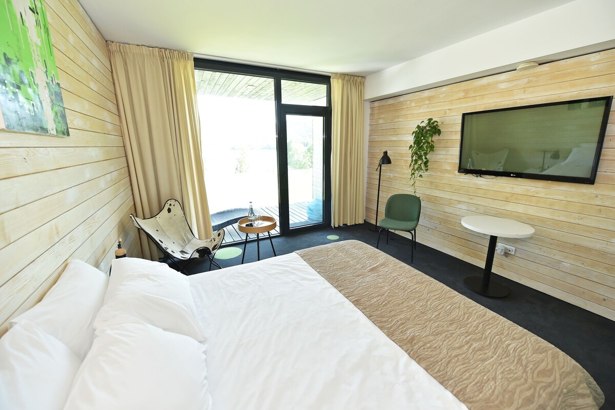 Habitación de hotel con cama, una mesita, una silla, televisión y un ventanal con cortinas.