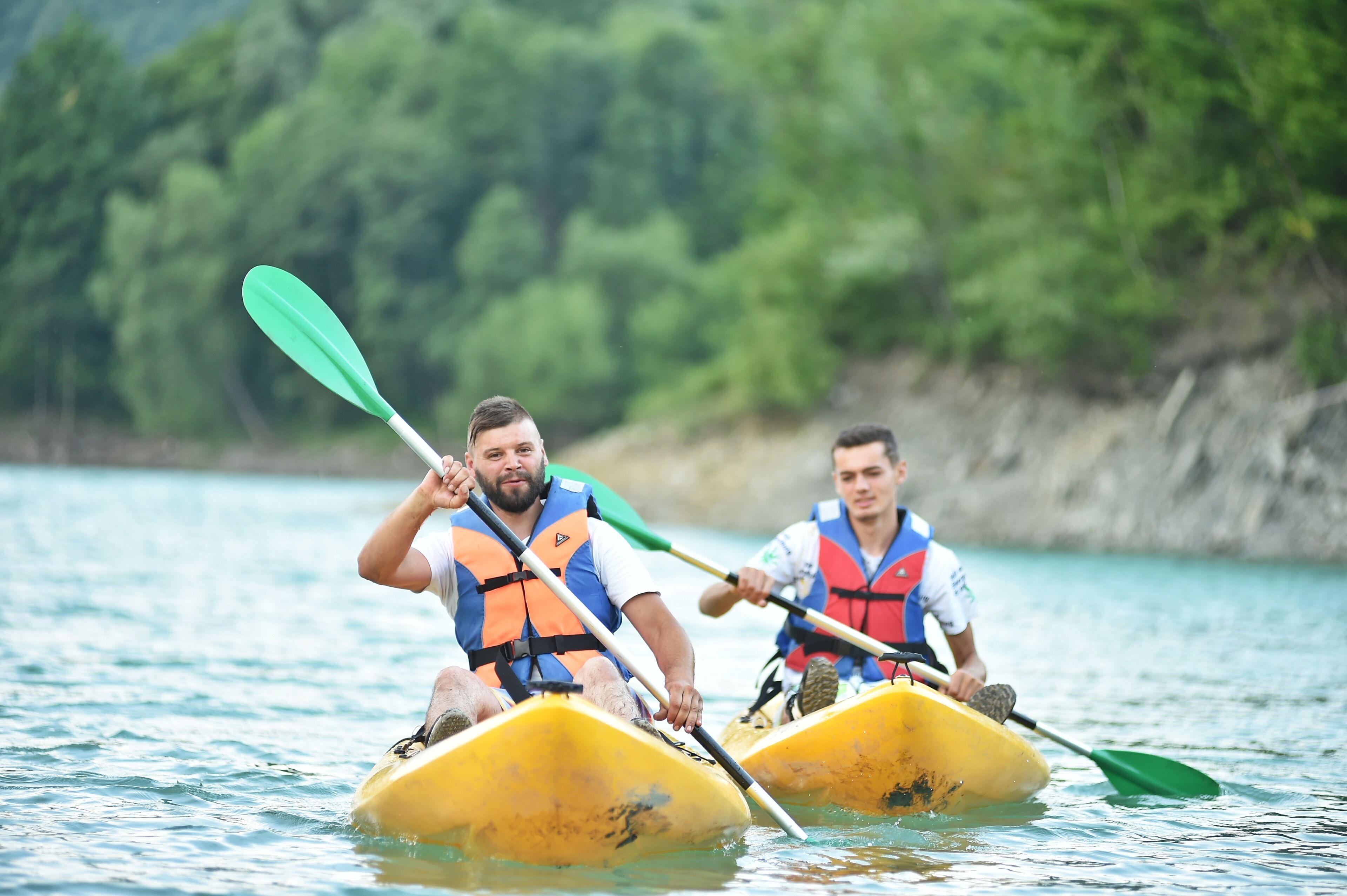 Two individuals kayaking on a calm body of water with a forested shoreline in the background.