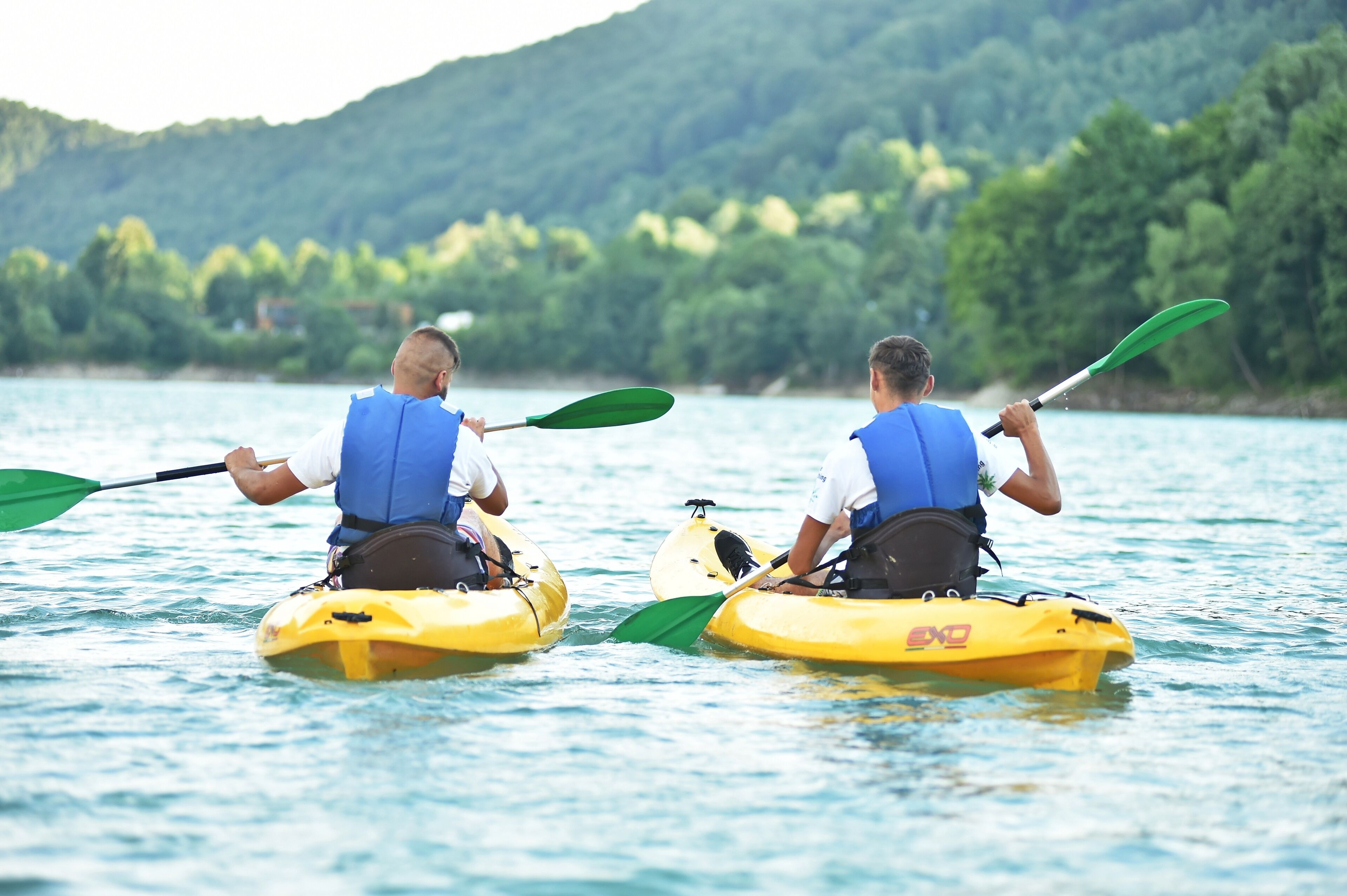 Deux personnes pratiquent le kayak sur un lac paisible, avec une colline boisée en arrière-plan.