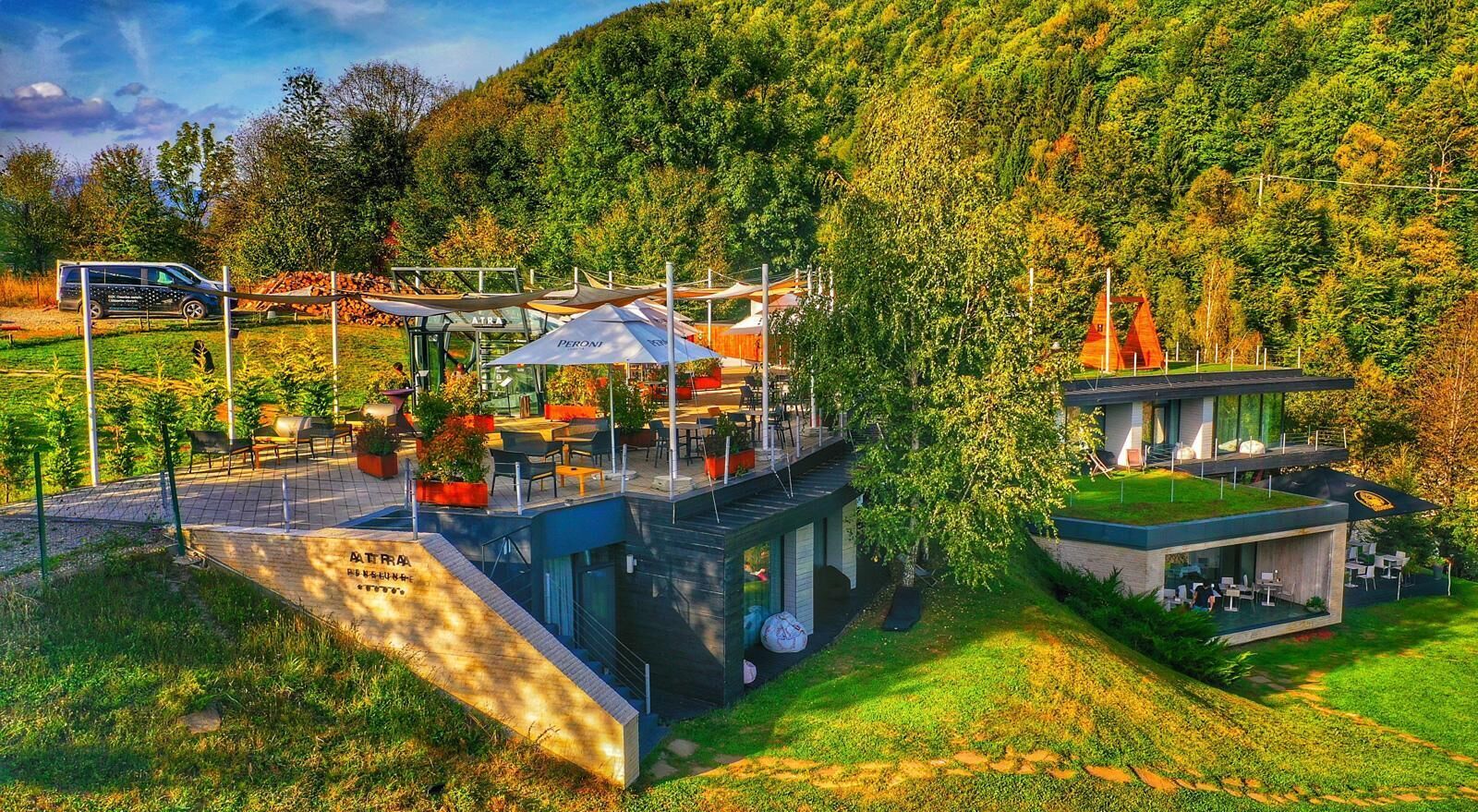 A modern outdoor café with tables and umbrellas, surrounded by lush greenery and a mountain backdrop.