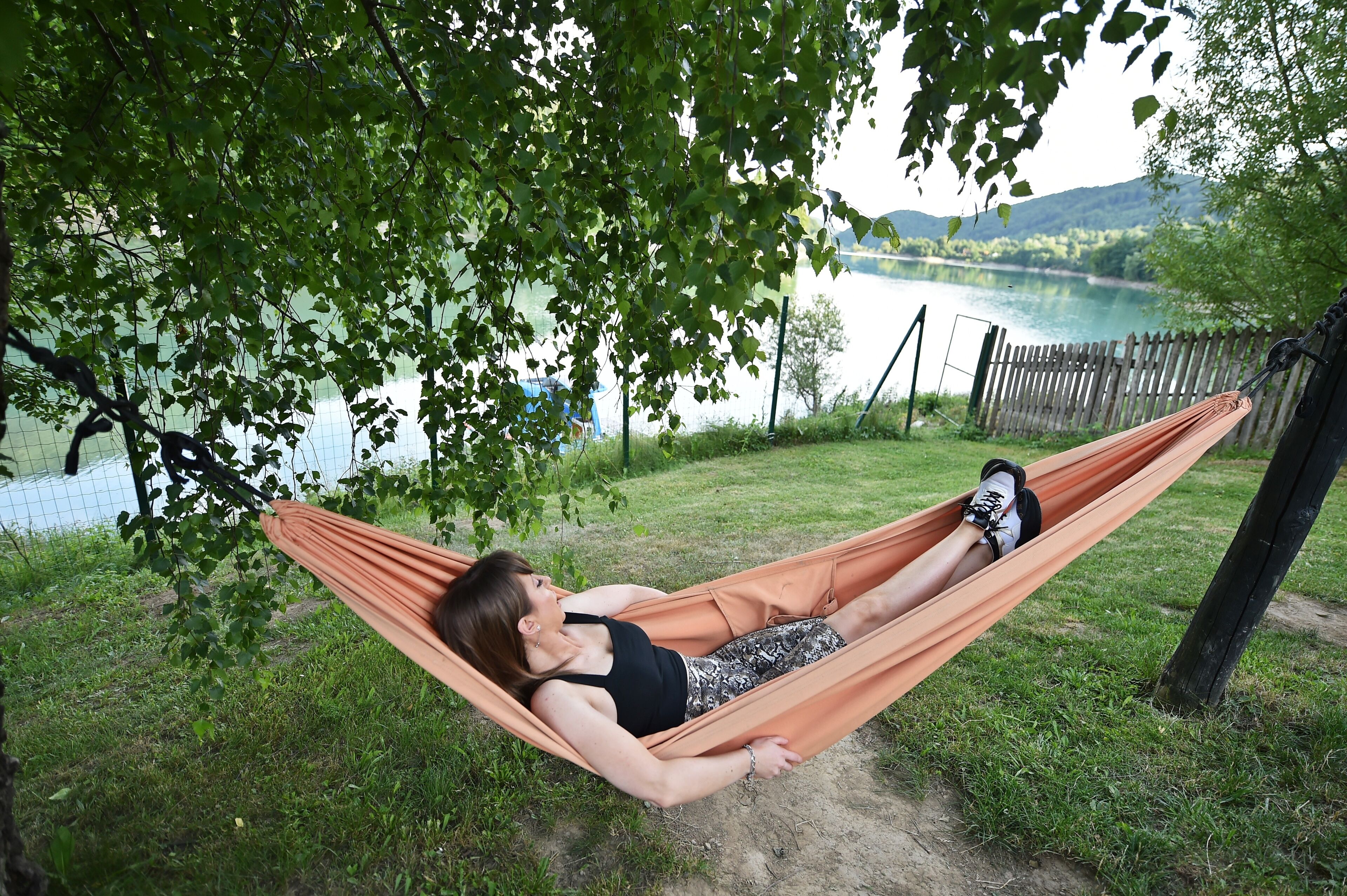 Une personne se détend dans un hamac au bord d’un lac, avec une clôture en bois et des arbres en arrière-plan.