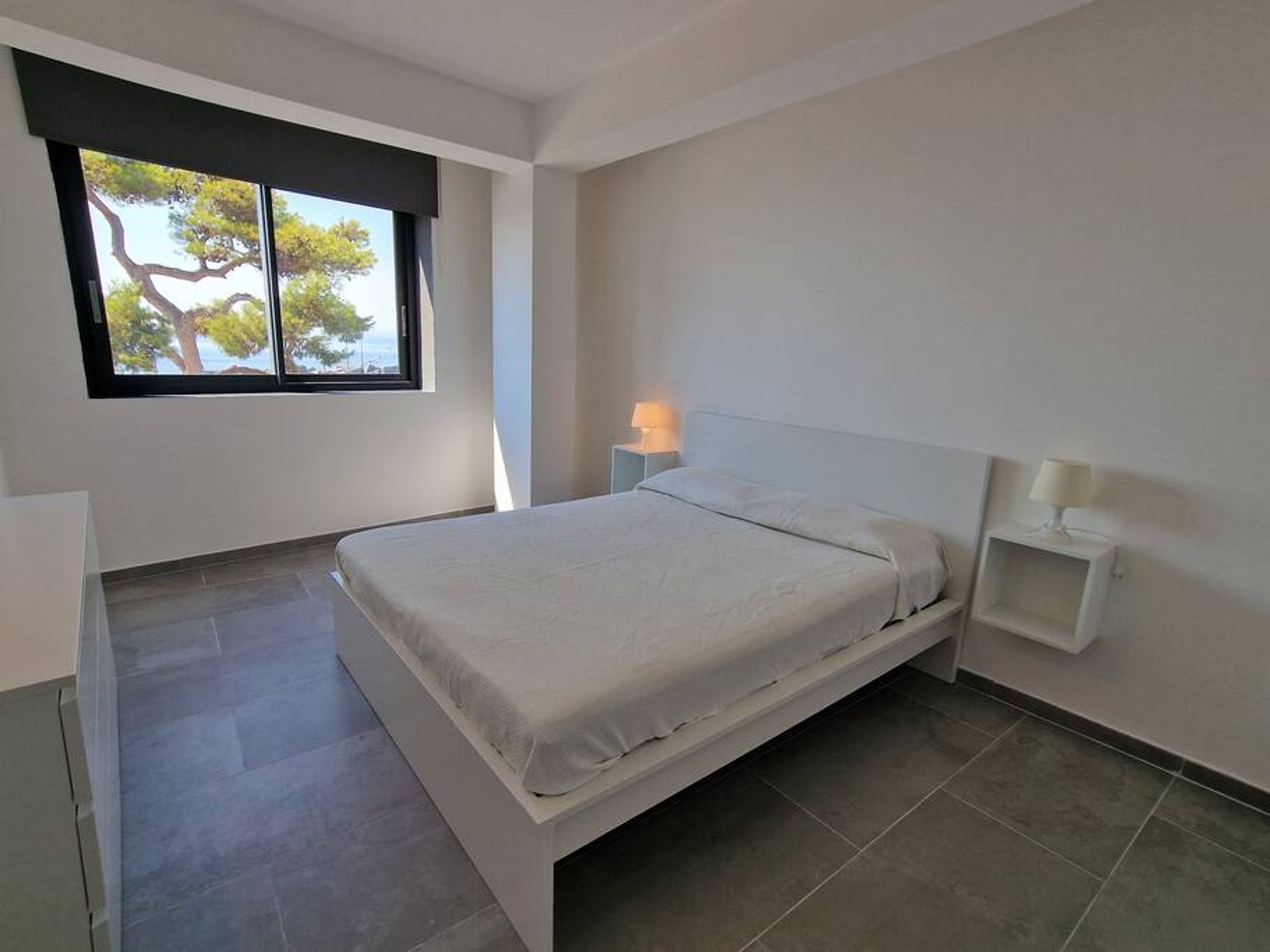 Quarto moderno com uma cama grande, uma mesa de cabeceira com abajur e uma janela com vista para árvores.